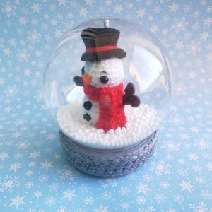 May include: A handcrafted snowman inside a clear snow globe. The snowman has a white body, a red scarf, and a black top hat. The base is gray and the globe sits on a blue snowflake background. A festive winter decoration.