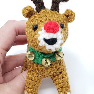 May include: A handmade crochet reindeer with a red nose, brown antlers, and a green collar with three gold bells. The reindeer has black eyes and white accents around its mouth. The body is a golden brown color.