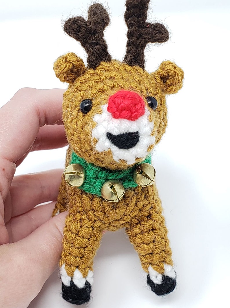 May include: A handmade crochet reindeer with a red nose, brown antlers, and a green collar with three gold bells. The reindeer has black eyes and white accents around its mouth. The body is a golden brown color.