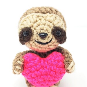May include: A crocheted sloth holding a pink heart. The sloth has brown eyes and a smiling mouth.