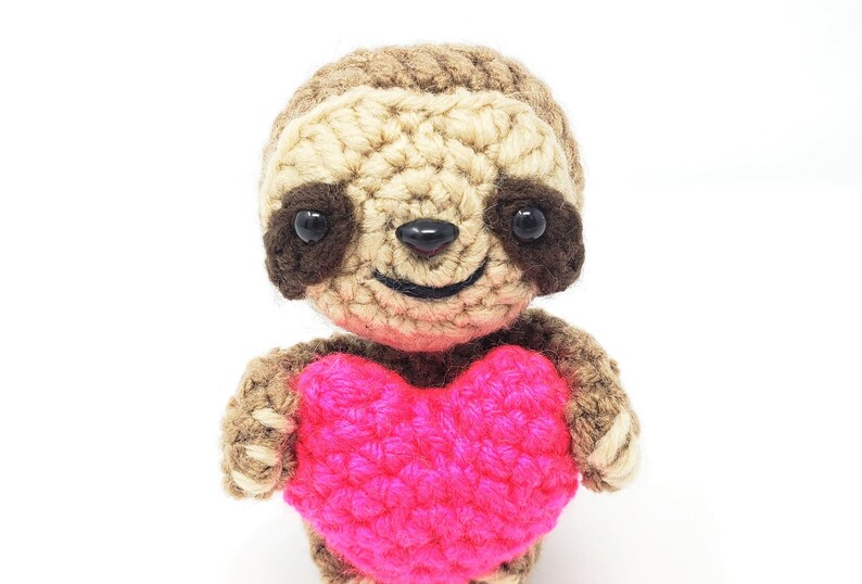 May include: A crocheted sloth holding a pink heart. The sloth has brown eyes and a smiling mouth.