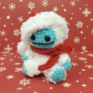 Op de afbeelding: Een handgemaakte gehaakte yeti-pop met een blauw gezicht en voeten, met een witte harige hoed en lichaam en een rode sjaal. De pop heeft zwarte ogen en een gestikte glimlach. De achtergrond is rood met witte sneeuwvlokpatronen.