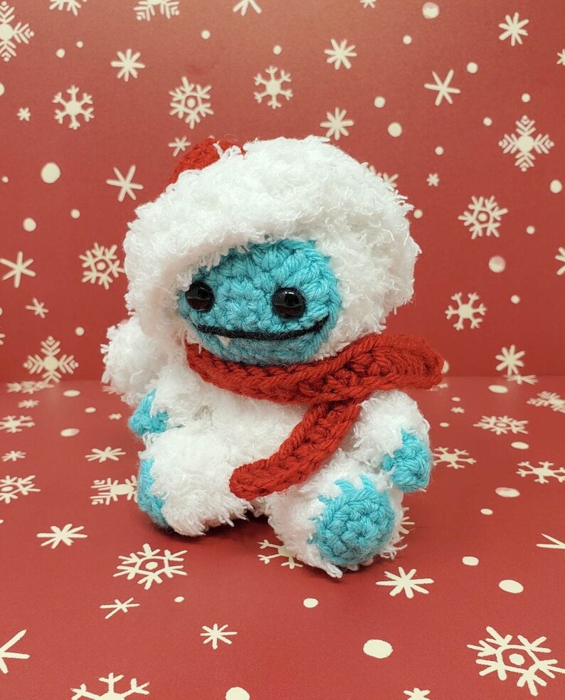 Op de afbeelding: Een handgemaakte gehaakte yeti-pop met een blauw gezicht en voeten, met een witte harige hoed en lichaam en een rode sjaal. De pop heeft zwarte ogen en een gestikte glimlach. De achtergrond is rood met witte sneeuwvlokpatronen.