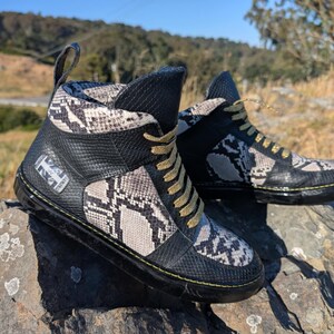 May include: High-top trainers featuring a snakeskin design with black panels. The footwear has gold laces and a black sole with yellow stitching. The shoes are displayed on a stone surface.