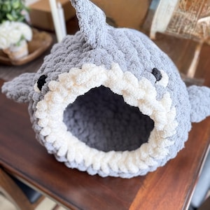 Peut inclure: Un lit pour animaux de compagnie en forme de requin, crocheté à la main, gris et blanc. Le lit présente un aileron de requin, des yeux noirs et une bouche blanche. L'intérieur est gris foncé.