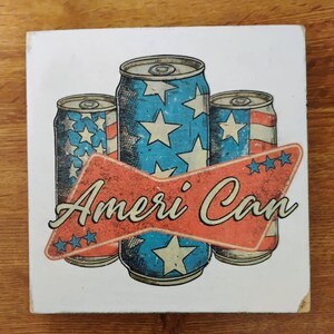 May include: A white square coaster featuring three stylized beverage cans with patriotic designs. The cans are blue with white stars and red and white stripes. The text "Ameri Can" is displayed on a red arrow-shaped banner.