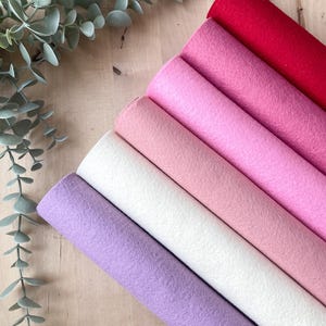 May include: A selection of colorful felt rolls in shades of red, pink, white, and purple, arranged diagonally. The felt is soft and textured, ideal for crafting projects. Green eucalyptus leaves are in the background.
