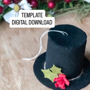 May include: A black felt top hat with a small brim, decorated with green felt holly leaves and a red paper flower. The hat has a white string attached. Text on the image reads: TEMPLATE DIGITAL DOWNLOAD.