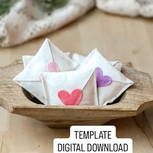 Op de afbeelding: Witte vilten envelopvormige decoraties met roze en paarse hartaccenten, weergegeven in een houten kom. De afbeelding bevat de tekst "TEMPLATE DIGITAL DOWNLOAD".