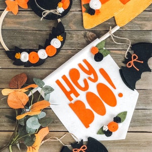 May include: Halloween-themed felt decorations, including a witch hat, candy corn, a banner with "Hey Boo!" text, a bat, and a wreath. The decorations feature orange, black, white, and green accents, perfect for seasonal decor.