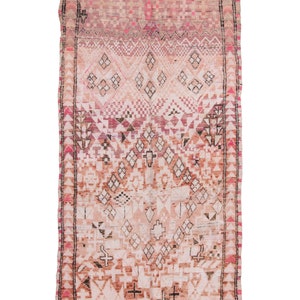 Puede incluir: Una alfombra marroquí vintage con un patrón geométrico rosa y blanco. La alfombra tiene un borde de flecos y está hecha de lana.