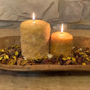 May include: Two lit, textured pillar candles in a wooden bowl filled with potpourri. The candles are different sizes and have a warm, golden hue. The bowl sits on a burlap surface, with a stone wall in the background.
