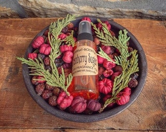 Pode incluir: Um pequeno frasco de vidro de óleo de fragrância Cranberry Sugar em uma tigela preta com bagas vermelhas e ramos verdes. O frasco tem um rótulo branco com texto preto que diz "Cranberry Sugar Fragrance Oil".