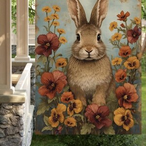 May include: A decorative garden flag featuring a detailed illustration of a brown rabbit surrounded by colorful flowers. The flowers are in shades of orange, yellow, and burgundy against a light blue background. The flag is designed for outdoor use.