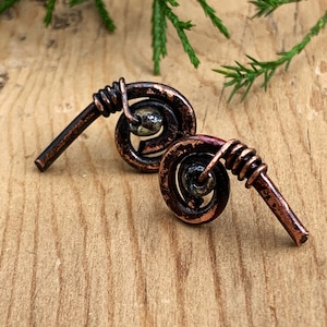 May include: A pair of handcrafted copper earrings. Each earring features a spiral design with a small, dark bead at the center. The copper has a rich, aged patina, and the earrings are set against a wooden background.
