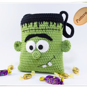 May include: Green crocheted Frankenstein monster trick or treat bag with black hair, white eyes, and a toothy grin. The bag has a black crocheted handle. The bag is surrounded by wrapped candies.