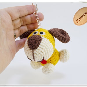 May include: A crocheted yellow and white dog keychain with brown ears and a red tongue. The keychain is attached to a silver chain.
