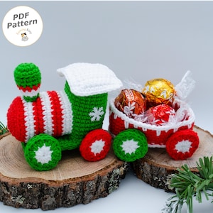 Può includere: Un trenino giocattolo all'uncinetto con locomotiva e vagone abbinato in rosso, verde e bianco. La locomotiva presenta un dettaglio a fiocco di neve bianco e il vagone contiene tre cioccolatini rotondi avvolti in carta dorata e rossa. Testo PDF Pattern.