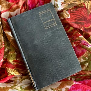 May include: A vintage book with a dark teal cover, titled "Shakespeare" from the "Academy Classics" series by Allyn and Bacon. The book rests on a floral patterned fabric with red, pink, and brown hues.