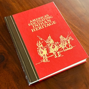 May include: A hardcover book titled "America's Fascinating Indian Heritage" with a red cover and gold illustrations of Native American figures on horseback. The spine is dark brown with gold accents.
