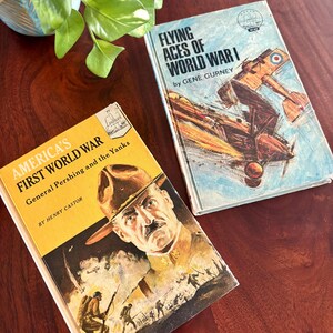 May include: Two vintage books on a wooden surface. One book, "America's First World War," features a portrait of General Pershing. The other, "Flying Aces of World War I," depicts a biplane. Both books have aged covers.