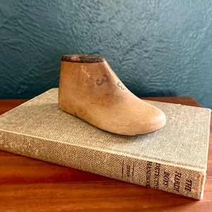 Puede incluir: Una horma de zapato de madera de color marrón claro con un borde superior marrón oscuro y el número 3 estampado en el lateral. La horma descansa sobre un libro de tapa dura gris claro titulado The Hardy Boys Detective Handbook con el nombre Dixon.