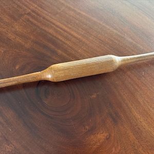 May include: A light brown wooden rolling pin featuring a central cylindrical barrel with a vertical ribbed texture and two smooth, tapered handles extending from each end. The wood grain is visible throughout the surface of the kitchen tool.