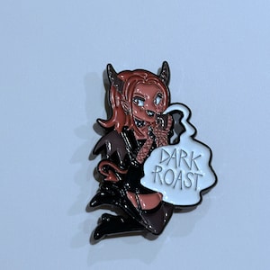 May include: A black and red enamel pin featuring a cartoon character with horns and a tail, holding a cup of coffee. The character is wearing a black dress and has a speech bubble that says "DARK ROAST".