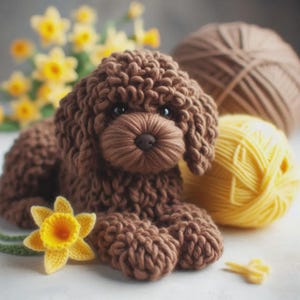May include: A handmade brown knitted dog with a detailed snout and curly fur. A yellow and white crocheted daffodil and a ball of yellow yarn are next to the dog. A ball of brown yarn is in the background.