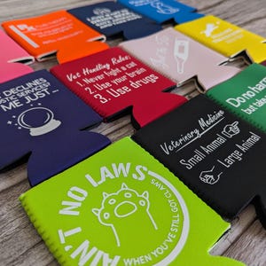 May include: A collection of colorful beverage can coolers with various printed phrases and graphics. The coolers are in shades of pink, orange, blue, red, green, yellow, and black. Some coolers have text like "Vet Handling Rules" and "No Laws".