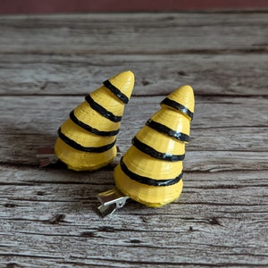Puede incluir: Dos pinzas para el pelo de color amarillo y negro con rayas en forma de cuernos. Las pinzas están hechas de fieltro y tienen un clip de metal en la parte posterior.