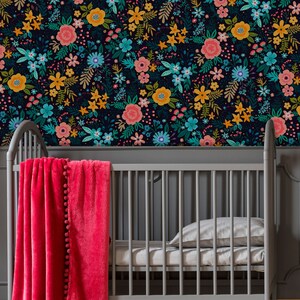 May include: A gray crib with a bright pink blanket adorned with pom-poms. The crib is set against a wall covered in a floral pattern featuring pink, yellow, and teal flowers on a dark background. A pillow and sheet are inside the crib.