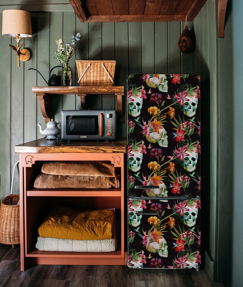 K&ouml;nnte beinhalten: Ein schwarzer K&uuml;hlschrank mit floralem und Totenkopf-Design. Die K&uuml;che verf&uuml;gt &uuml;ber eine Mikrowelle, Regale und einen Schrank. Das Farbschema umfasst Gr&uuml;n-, Orange- und Braunt&ouml;ne und schafft eine einzigartige und k&uuml;nstlerische &Auml;sthetik.