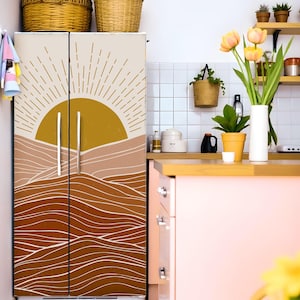 K&ouml;nnte beinhalten: Ein K&uuml;hlschrank mit dekorativer Vinylfolie, die eine Sonne und Landschaft in Gold-, Beige- und Braunt&ouml;nen zeigt. Die K&uuml;chenumgebung umfasst K&ouml;rbe, Pflanzen und einen rosa Schrank.