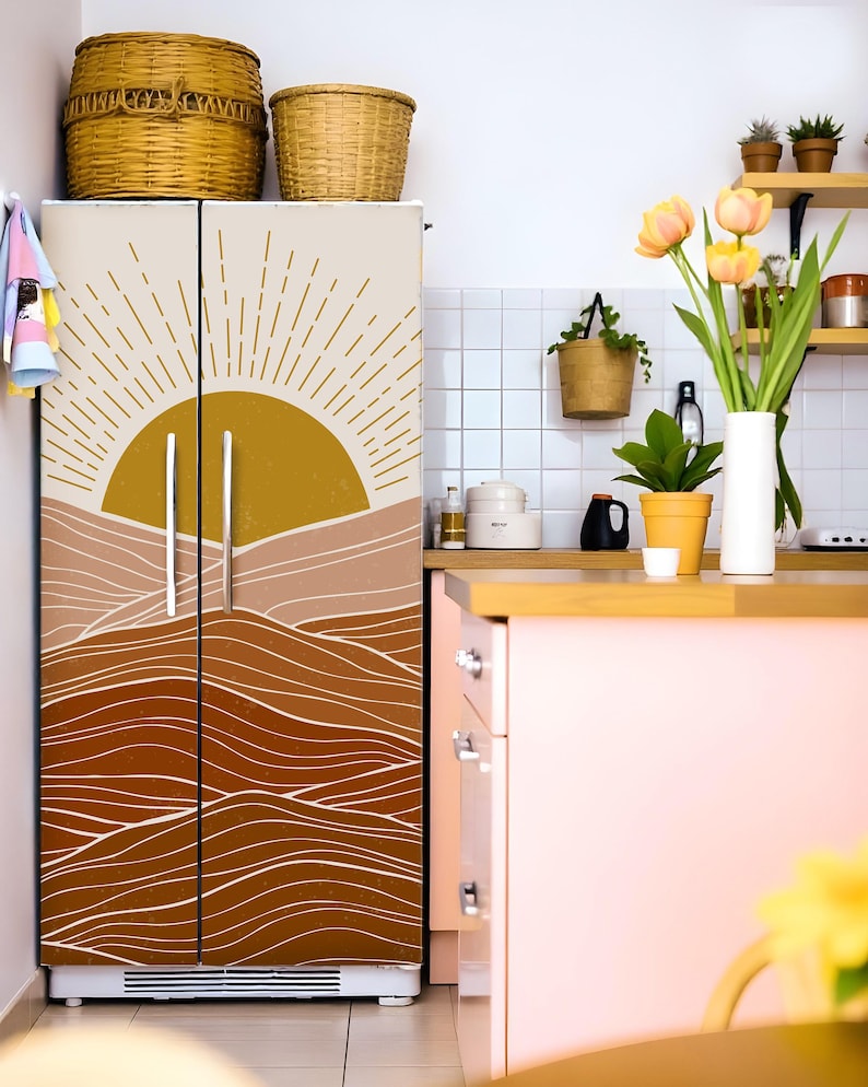 K&ouml;nnte beinhalten: Ein K&uuml;hlschrank mit dekorativer Vinylfolie, die eine Sonne und Landschaft in Gold-, Beige- und Braunt&ouml;nen zeigt. Die K&uuml;chenumgebung umfasst K&ouml;rbe, Pflanzen und einen rosa Schrank.