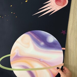 Pode incluir: Um planeta roxo e laranja com um anel verde ao redor contra um fundo azul escuro com um cometa rosa e estrelas amarelas.