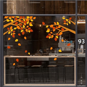 Könnte beinhalten: Ein Fensteraufkleber-Set mit zwei braunen Baumzweigen und verstreuten Herbstblättern in Orange-, Gelb- und Rottönen. Die Blätter wirken wie herabfallend und erzeugen eine saisonale Dekoration auf der Glasfläche eines Schaufensters.