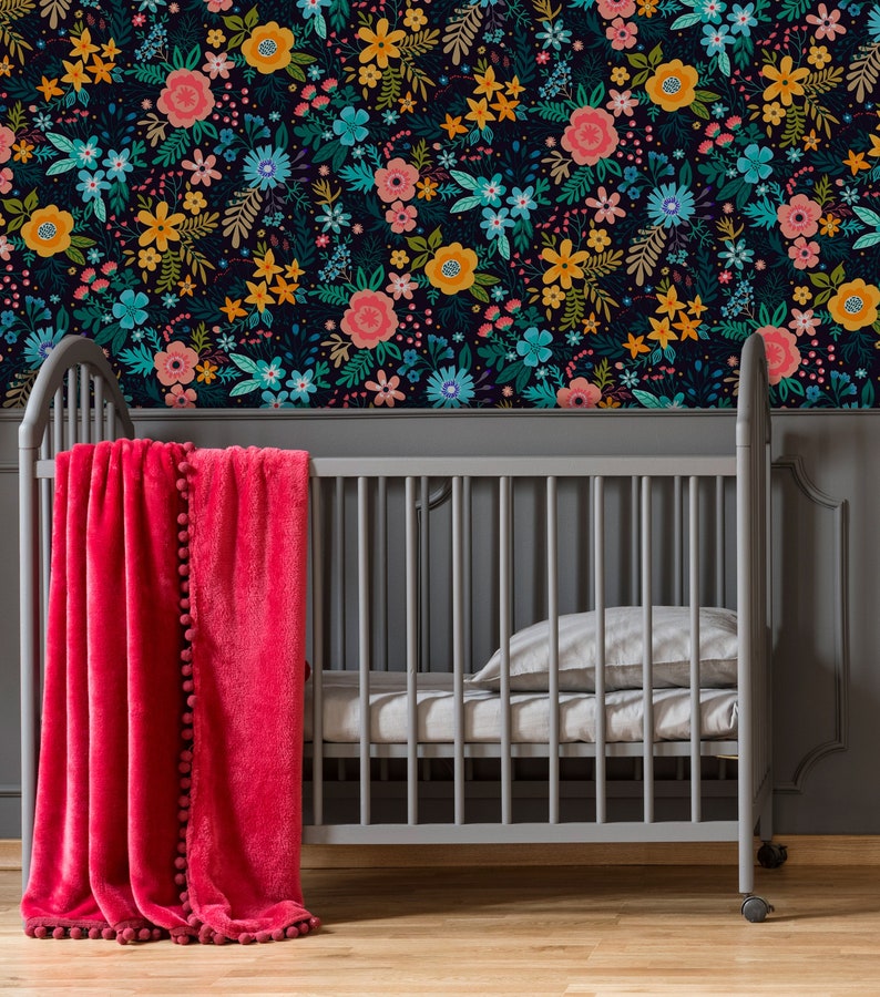 May include: A gray crib with a bright pink blanket adorned with pom-poms. The crib is set against a wall covered in a floral pattern featuring pink, yellow, and teal flowers on a dark background. A pillow and sheet are inside the crib.