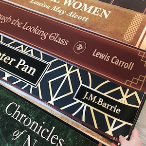 K&ouml;nnte beinhalten: Ein Stapel von f&uuml;nf B&uuml;chern mit dekorativen R&uuml;ckseiten. Das oberste Buch tr&auml;gt den Titel "Little Women" von Louisa May Alcott. Das zweite Buch tr&auml;gt den Titel "Through the Looking Glass" von Lewis Carroll. Das dritte Buch tr&auml;gt den Titel "Peter Pan" von J.M. Barrie. Das vierte Buch tr&auml;gt den Titel "Chronicles of Narnia".