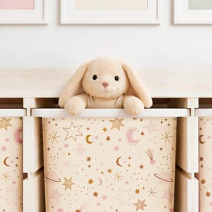 May include: A beige plush bunny toy peeks over the edge of a storage bin. The bin is decorated with a celestial pattern of stars, moons, and planets in gold and pink hues. The storage bin is part of a set.