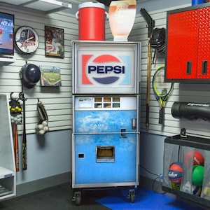 May include: A vintage Pepsi vending machine with the Pepsi logo in red, white, and blue. The machine is blue and has the words "Served in Cans" on it. A red cooler and a clear plastic container sit on top. The image is in a garage setting.