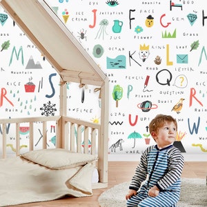 May include: A white wall with colorful letters of the alphabet, each letter is paired with a word and an illustration. The letters are in a playful, hand-drawn style. A young child is sitting in front of the wall, looking up at the letters.