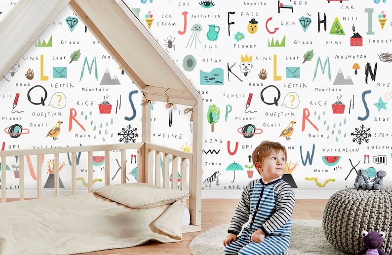 May include: A white wall with colorful letters of the alphabet, each letter is paired with a word and an illustration. The letters are in a playful, hand-drawn style. A young child is sitting in front of the wall, looking up at the letters.