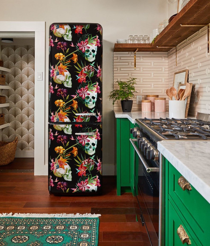 K&ouml;nnte beinhalten: Ein schwarzer K&uuml;hlschrank mit einem farbenfrohen Blumen- und Totenkopfmuster. Das Design zeigt wei&szlig;e Totenk&ouml;pfe, rote, rosa und gelbe Blumen. Der K&uuml;hlschrank steht in einer K&uuml;che mit gr&uuml;nen Schr&auml;nken und Holzboden.