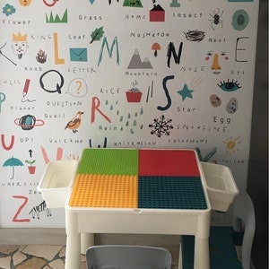 May include: White plastic table with a colorful square building block top. The table has two small white plastic trays on the sides. The table is in front of a wall with a colorful alphabet chart.
