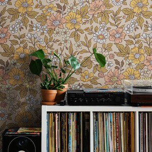 May include: A white bookshelf with multiple shelves filled with vinyl records. A black stereo system sits on top of the bookshelf. A potted plant with green leaves sits on the shelf to the left of the stereo system. The wall behind the bookshelf is covered in floral wallpaper with a brown background and yellow, pink, and white flowers.
