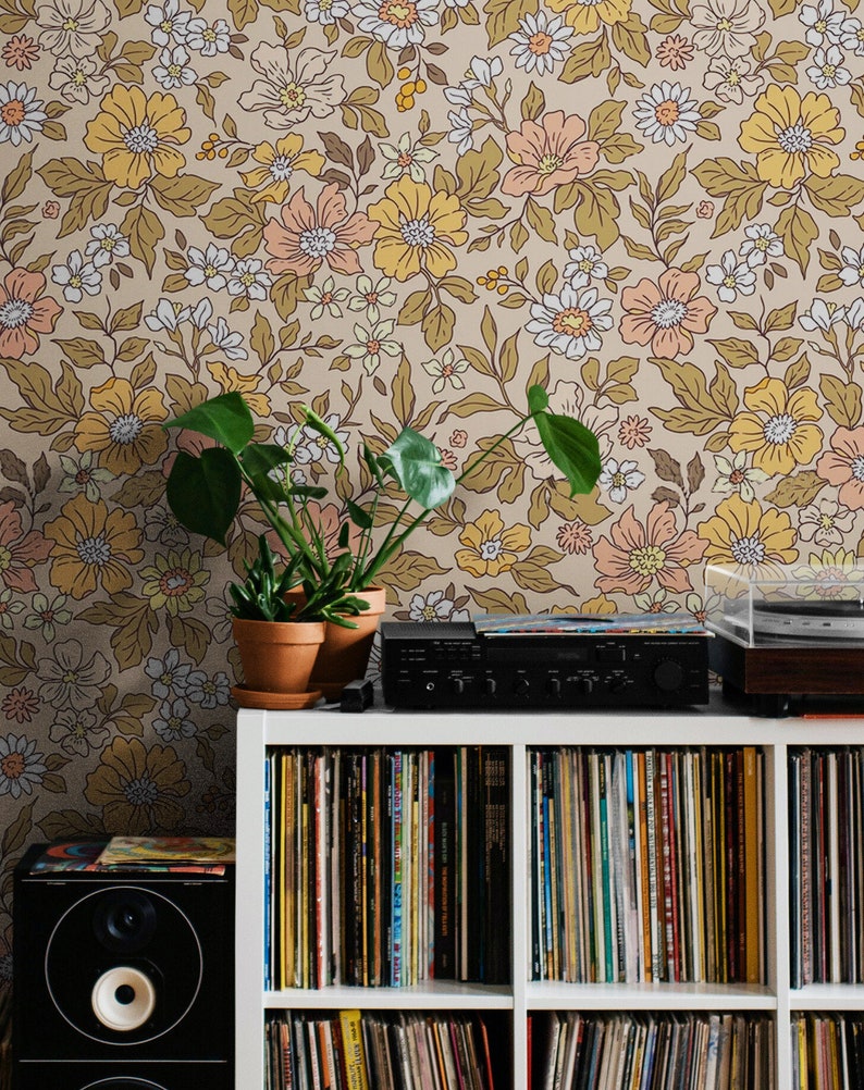 May include: A white bookshelf with multiple shelves filled with vinyl records. A black stereo system sits on top of the bookshelf. A potted plant with green leaves sits on the shelf to the left of the stereo system. The wall behind the bookshelf is covered in floral wallpaper with a brown background and yellow, pink, and white flowers.