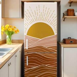 K&ouml;nnte beinhalten: Ein K&uuml;hlschrank mit einer dekorativen Vinylfolie mit einem Sonnen- und Landschaftsdesign. Das Kunstwerk zeigt eine goldene Sonne mit Strahlenlinien und geschichteten braunen und beigen H&uuml;geln. Der K&uuml;hlschrank hat wei&szlig;e Griffe.
