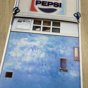 May include: A vintage Pepsi-Cola vending machine print. The top features the Pepsi logo in red, white, and blue. The machine's body is blue with the words "Served in Cans." Scissors are on either side of the print.