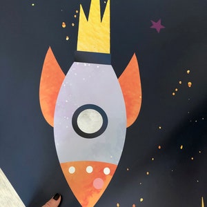 Pode incluir: Um foguete roxo e laranja com uma chama amarela em um fundo azul escuro com estrelas douradas e brancas.